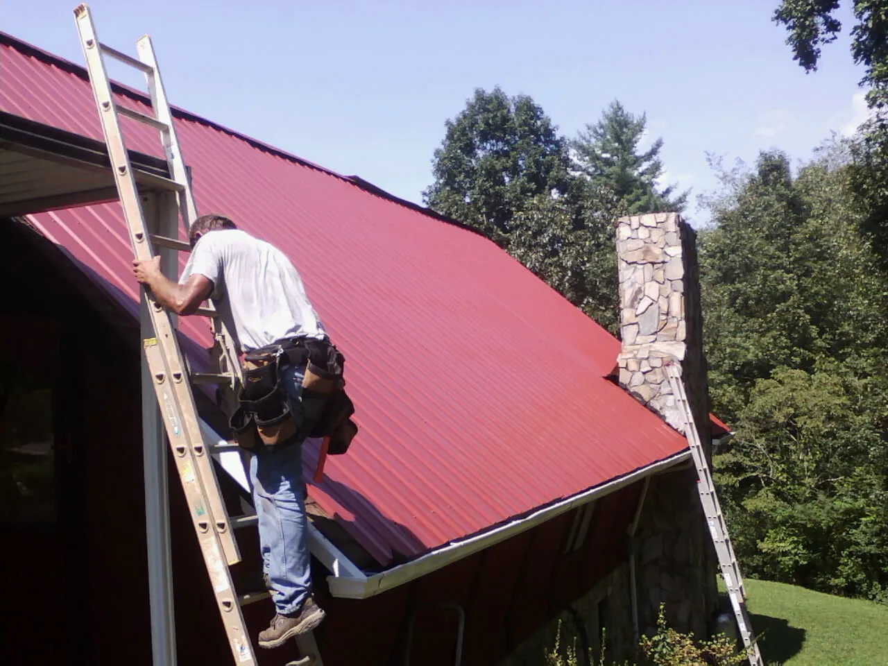Roofer on a ladder beside a red metal roof and stone chimney — Soffit & Fascia Repair by Roofing Brothers in Millstone Township